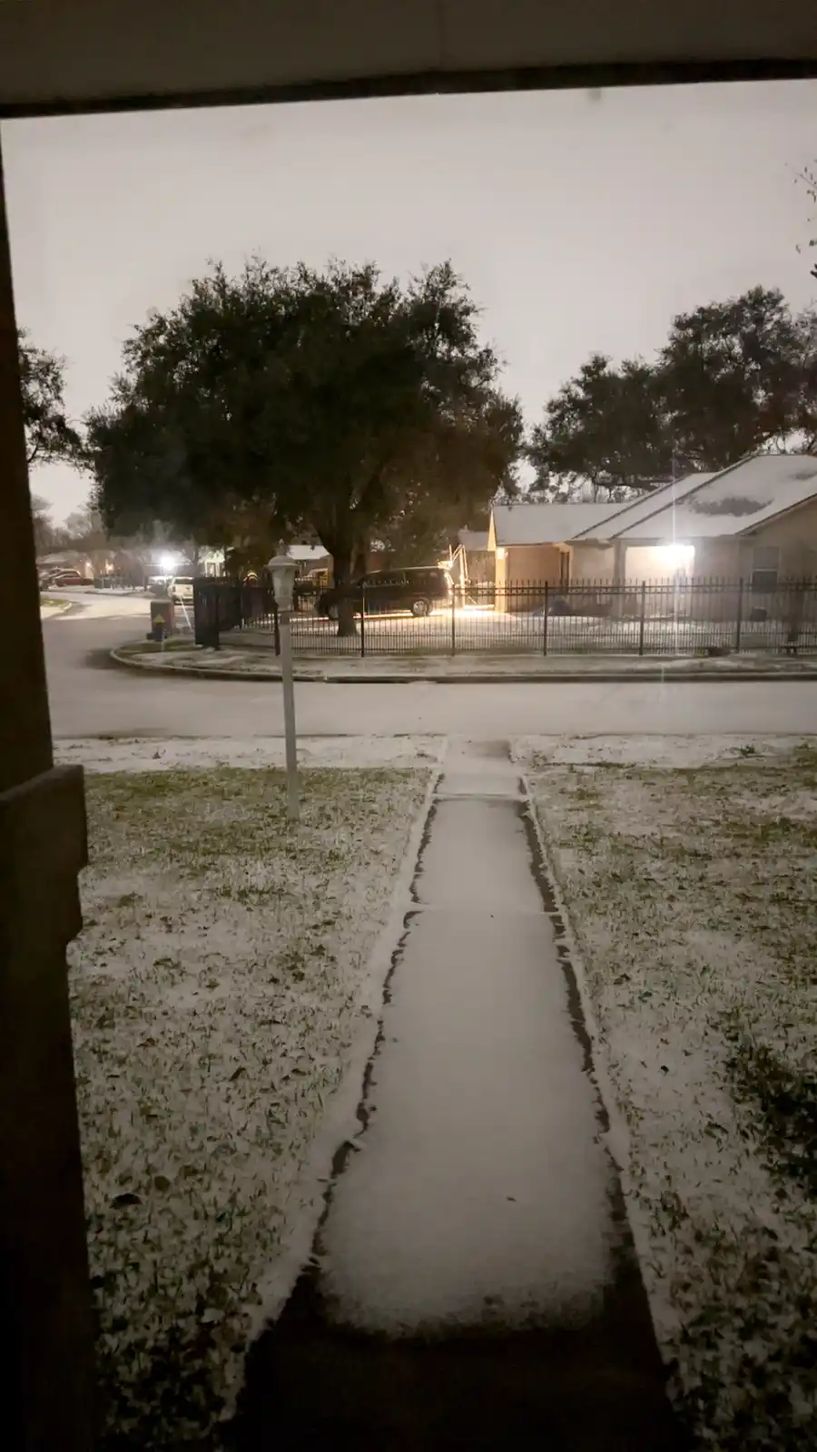A snowy suburban street scene viewed from a porch, showing a snow-covered sidewalk and grass, with houses and trees in the…