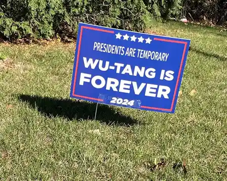 A blue political yard sign with red trim and white text stands in a grassy yard, featuring five stars at the top. The text…