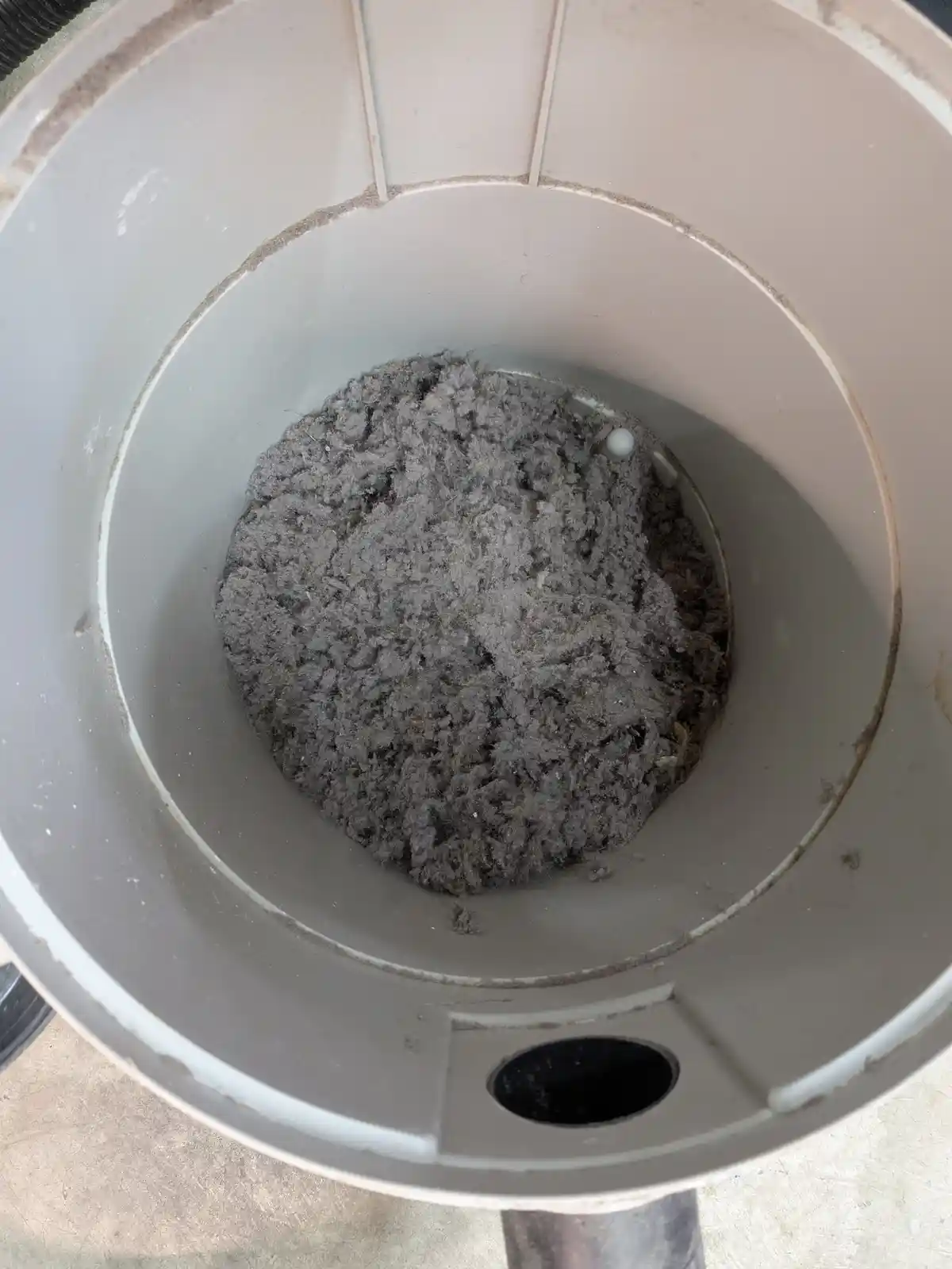 A top-down view of a dirty vacuum cleaner dustbin filled with a large, gray, fibrous accumulation of dust and debris. The…