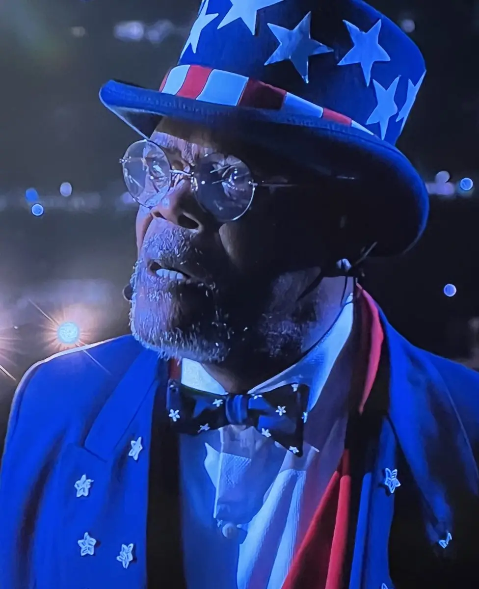 A man wearing a patriotic Uncle Sam-style hat with stars and stripes, round glasses, and a blue suit with star pins, looks…