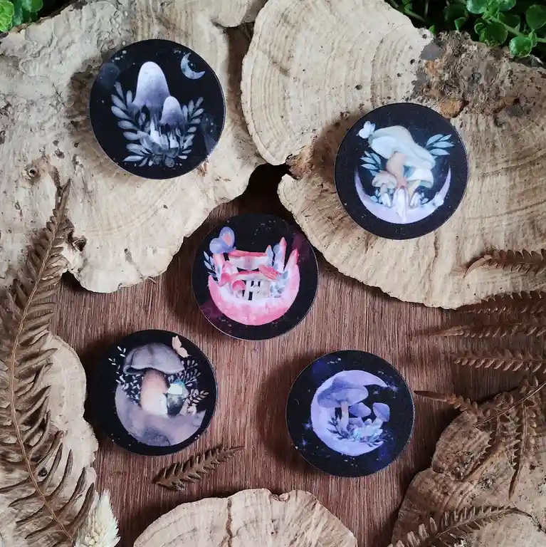 Five circular decorative items with mushroom-themed illustrations are arranged on rustic wood slices and dried ferns. Each…