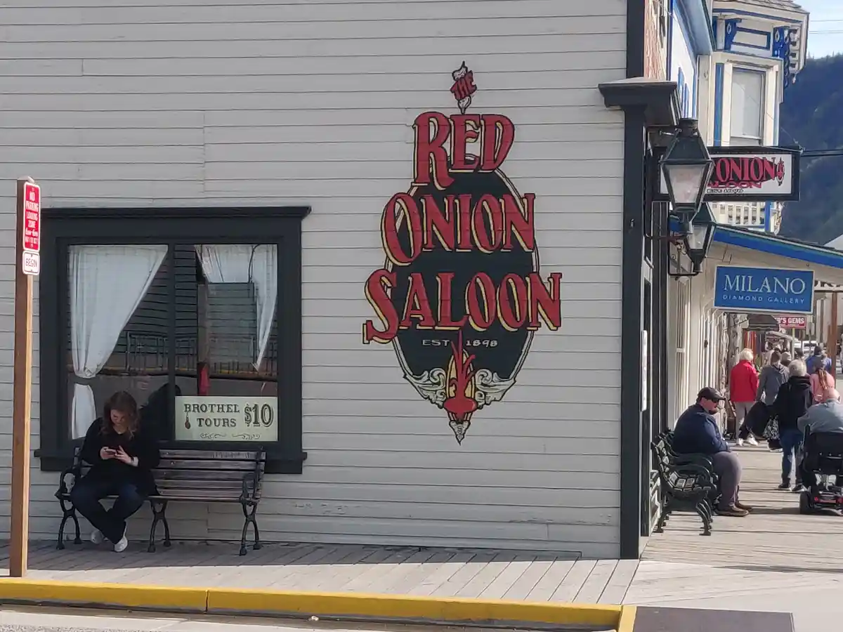 A woman sits on a bench outside The Red Onion Saloon, a historic building with a large red and black sign that reads 'EST…