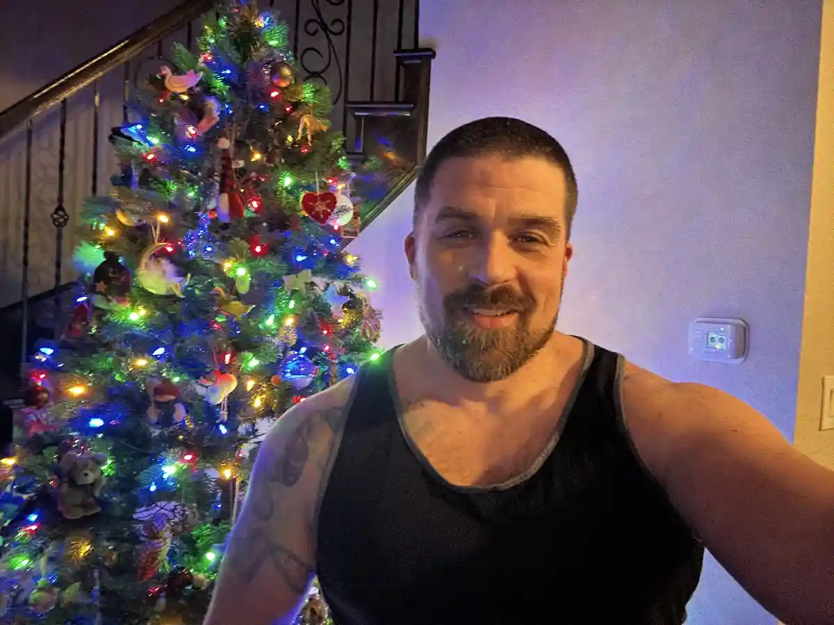 A man with a beard and tattoos, wearing a black tank top, takes a selfie in front of a brightly lit, decorated Christmas…