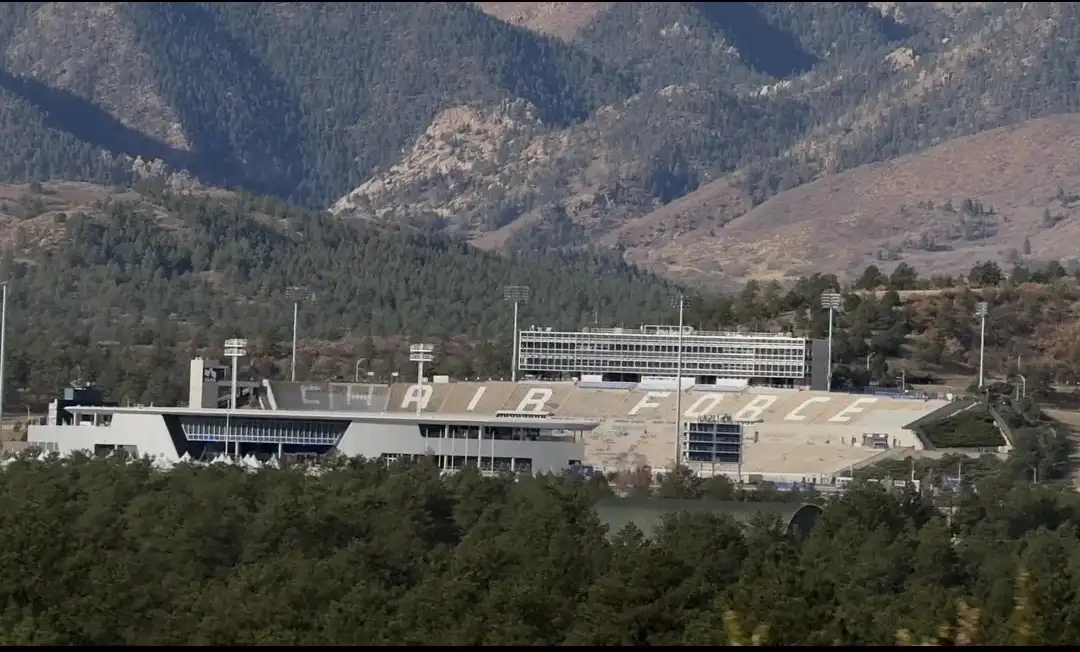 An aerial view of a large stadium nestled in a mountainous, forested area. The stadium's tiered seating has the words 'AIR…