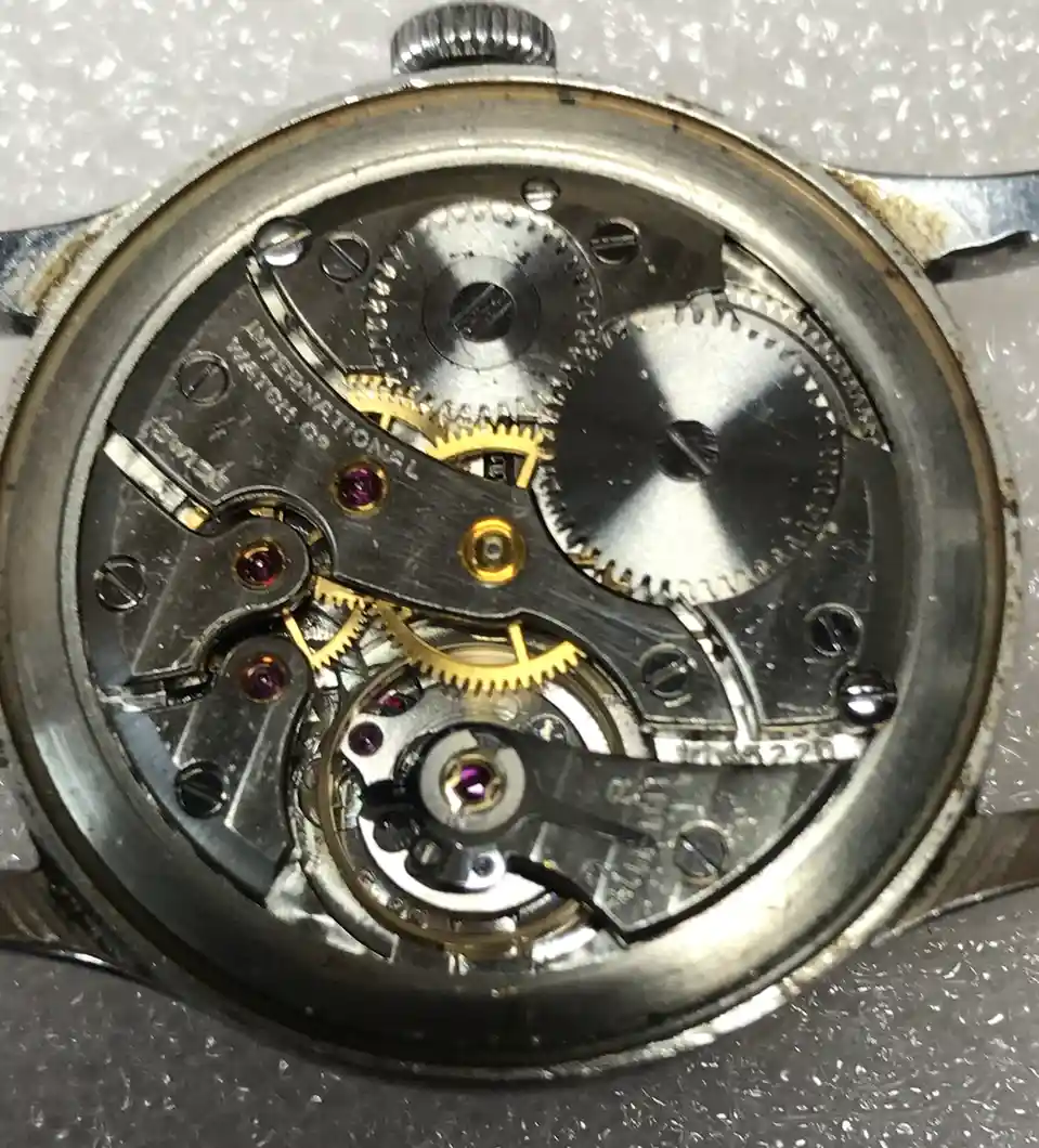 A close-up photo of the exposed internal mechanical movement of an old, worn pocket watch, showing gears, screws, and ruby…