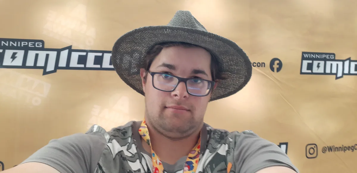 A man wearing glasses and a wide-brimmed hat takes a selfie in front of a Winnipeg Comic Con backdrop. He has a neutral…