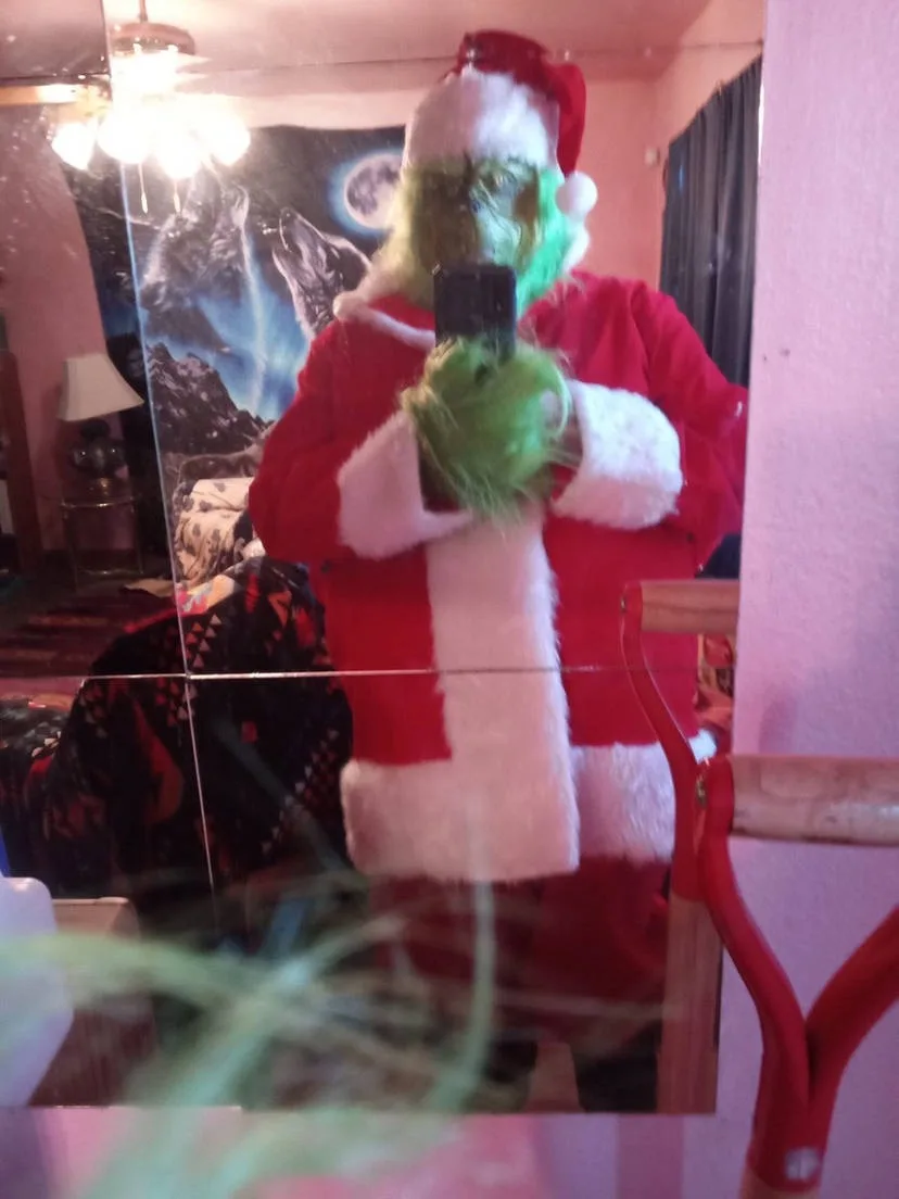 A person wearing a green Grinch mask and a red Santa suit with white trim takes a mirror selfie, holding a phone. The…
