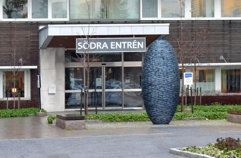 An exterior shot of a modern building entrance labeled 'SÖDRA ENTRÉN' with glass doors and a large, dark blue, egg-shaped…