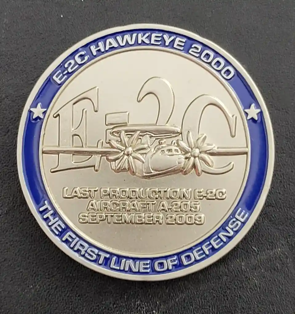 A commemorative challenge coin features an embossed E-2C Hawkeye aircraft with large 'E-2C' lettering behind it. The blue…