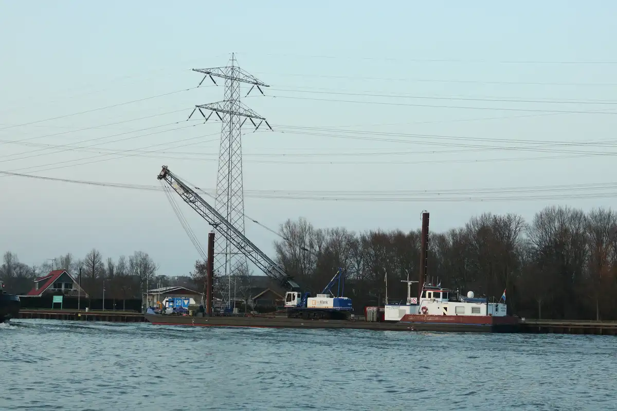 A large crane mounted on a barge is positioned directly under a tall electricity pylon, with its boom extended upward. The…