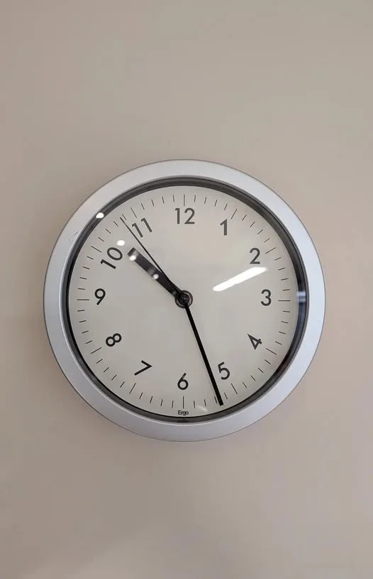 A close-up photo of a round, silver-framed wall clock mounted on a plain beige wall. The clock face is white with black…