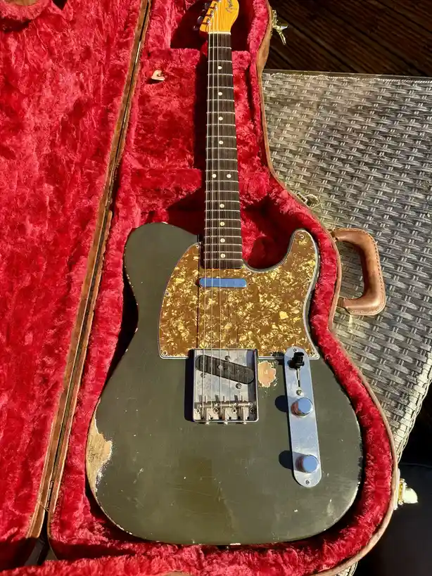 A vintage-style electric guitar with a dark green body and gold pickguard rests inside an open, red velvet-lined case. The…