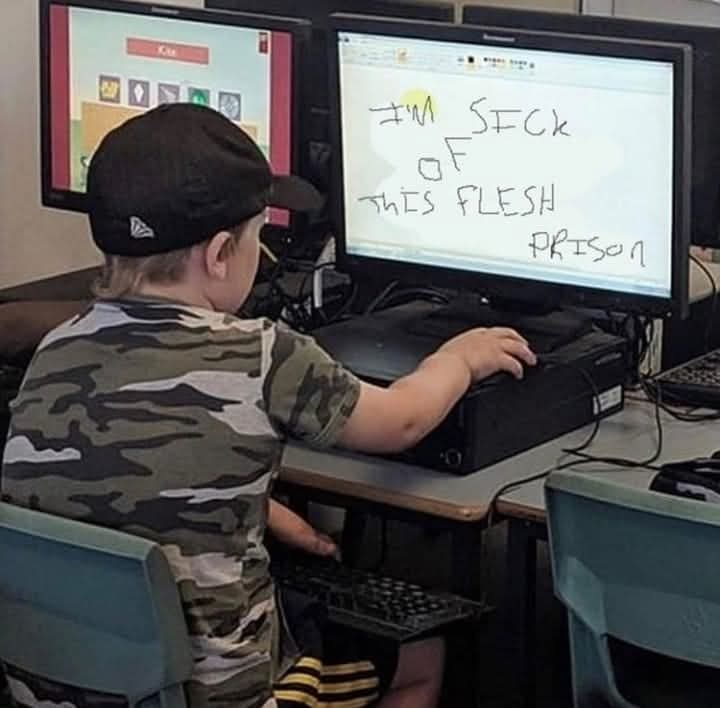 A young child in a camouflage shirt and black cap sits at a desk, viewed from behind, using a computer. The child's right…
