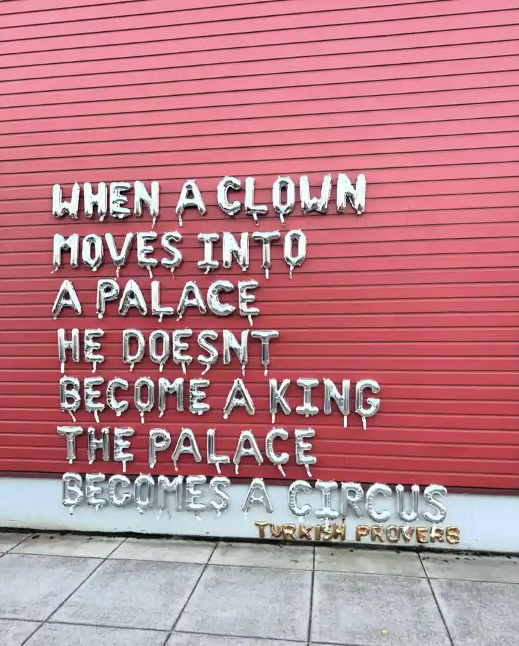 Silver foil balloon letters spell out a humorous proverb on a red corrugated metal wall, with smaller gold letters below…