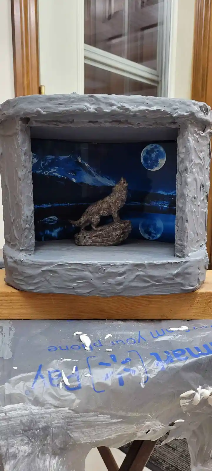A handmade diorama with a gray, textured frame resembling stone, containing a wolf figurine howling at a blue moon against a…