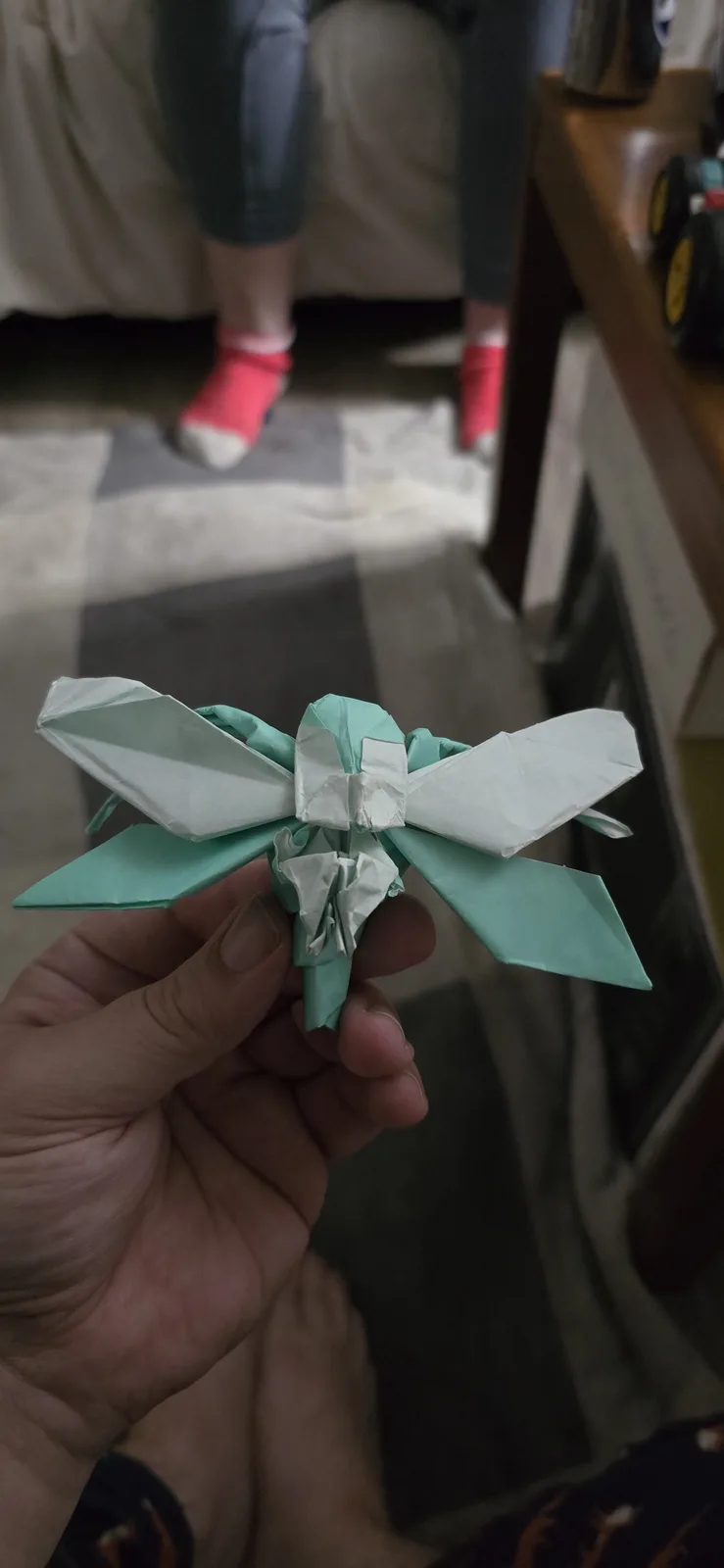 A hand holds a small origami insect made from light green and white paper, featuring large wings and segmented body. The…
