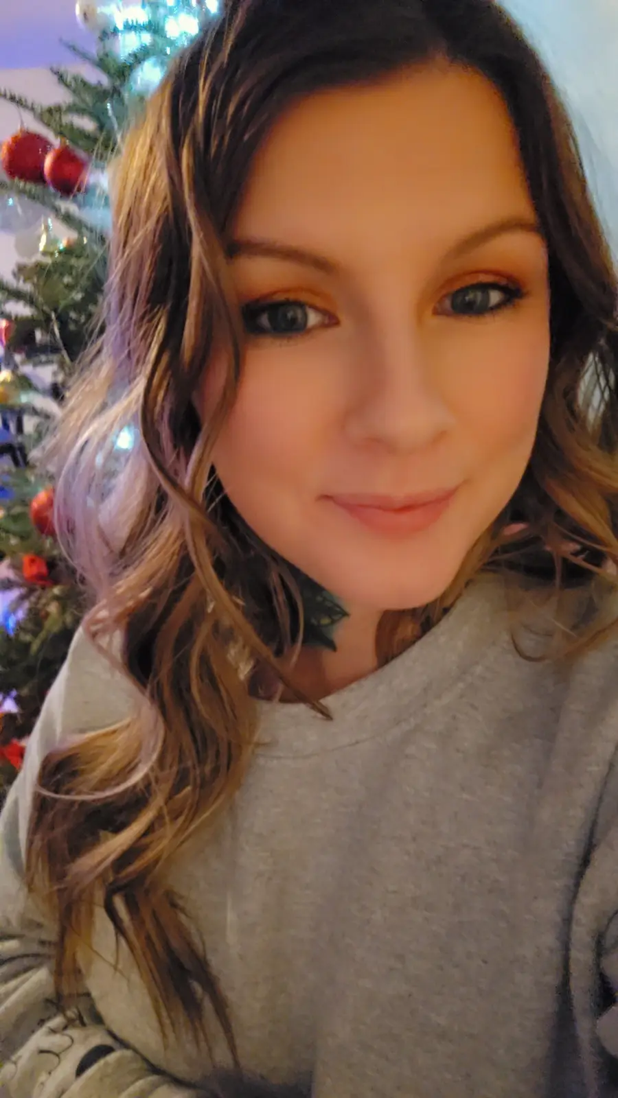 A selfie of a woman with long brown hair and a grey sweatshirt, smiling slightly in front of a decorated Christmas tree with…