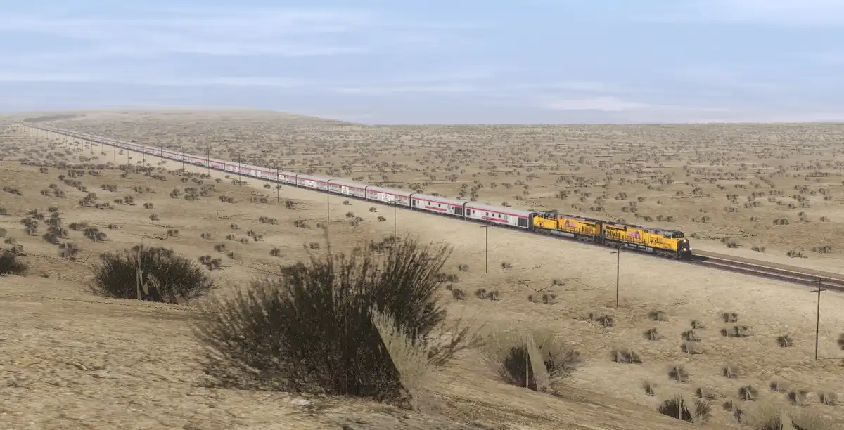 A long passenger train with yellow locomotives travels across a vast, arid desert landscape under a pale blue sky. The train…