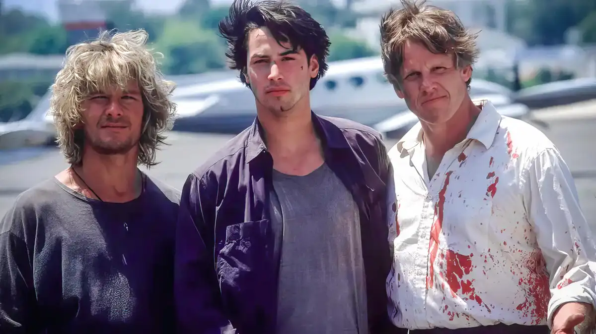 Three men stand side-by-side outdoors, with the man on the right wearing a white shirt splattered with red stains resembling…