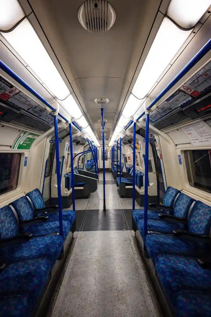 An empty, brightly lit subway train carriage with blue patterned seats, blue poles, and overhead maps, overlaid with the…