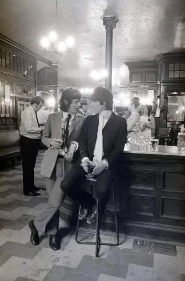 A black-and-white photo shows two men, identified as Mick Jagger and Keith Richards of the Rolling Stones, in a vintage bar…