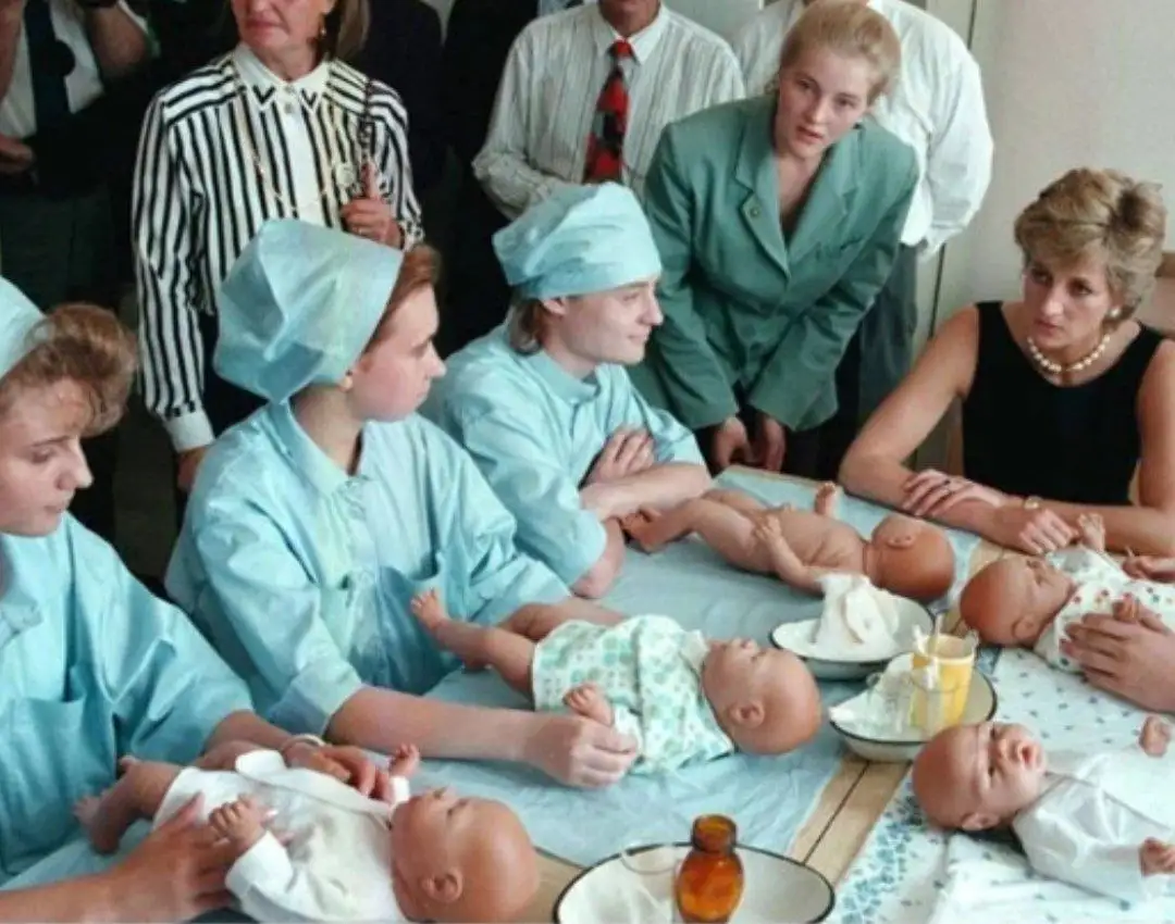 A historical photo shows Princess Diana observing nurses in light blue scrubs practicing infant care on baby dolls laid out…