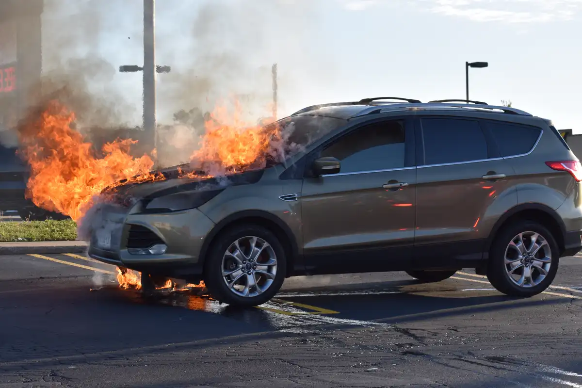 A silver SUV is engulfed in bright orange flames and thick smoke, primarily concentrated on the front hood and bumper area…
