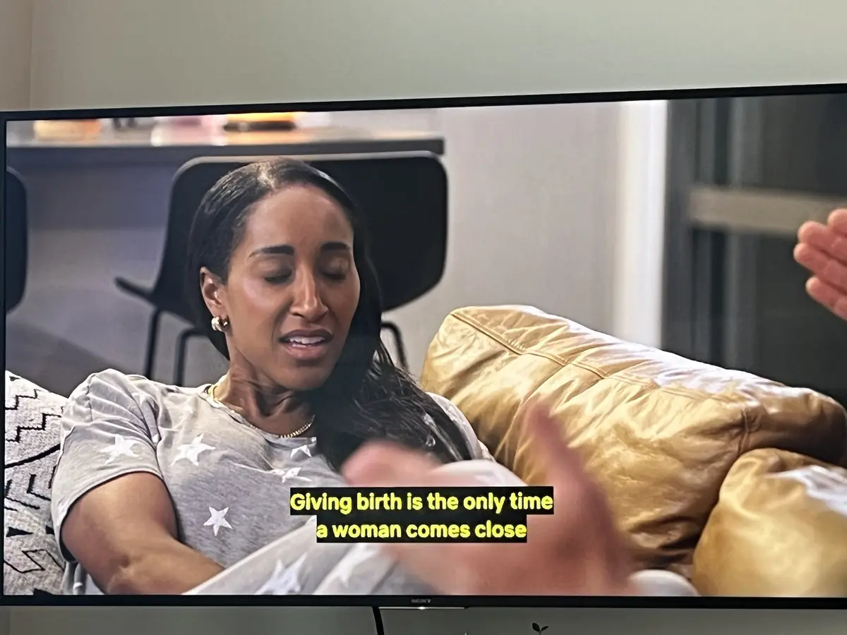 A television screen shows a woman with dark hair and a grey star-patterned shirt sitting on a gold-colored couch, looking…