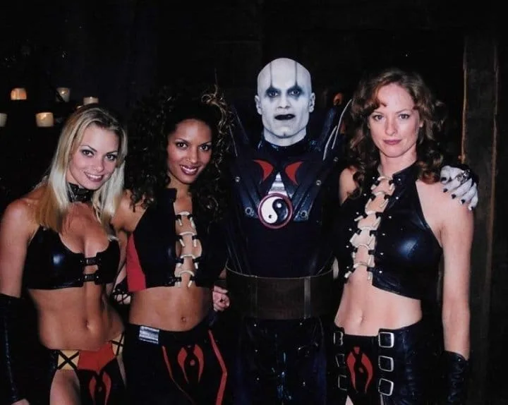 Four people pose together in a dark room with candles; a bald man in a black leather costume with a yin-yang symbol on his…