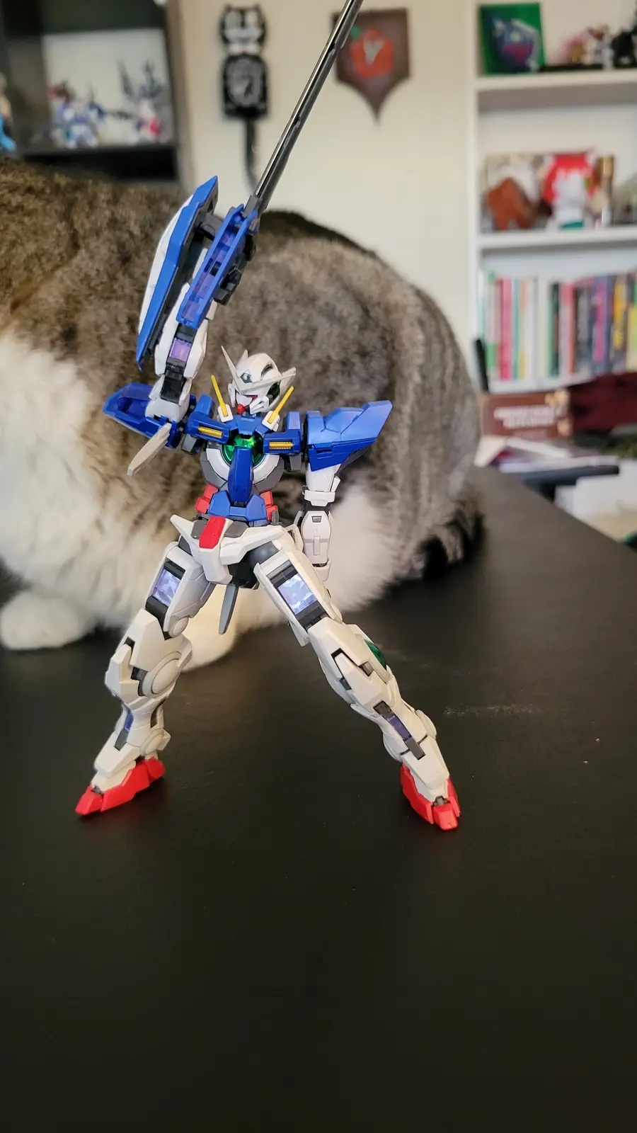 A blue, white, and red Gundam model robot stands on a dark table in a heroic pose, holding a long black sword. Behind it, a…