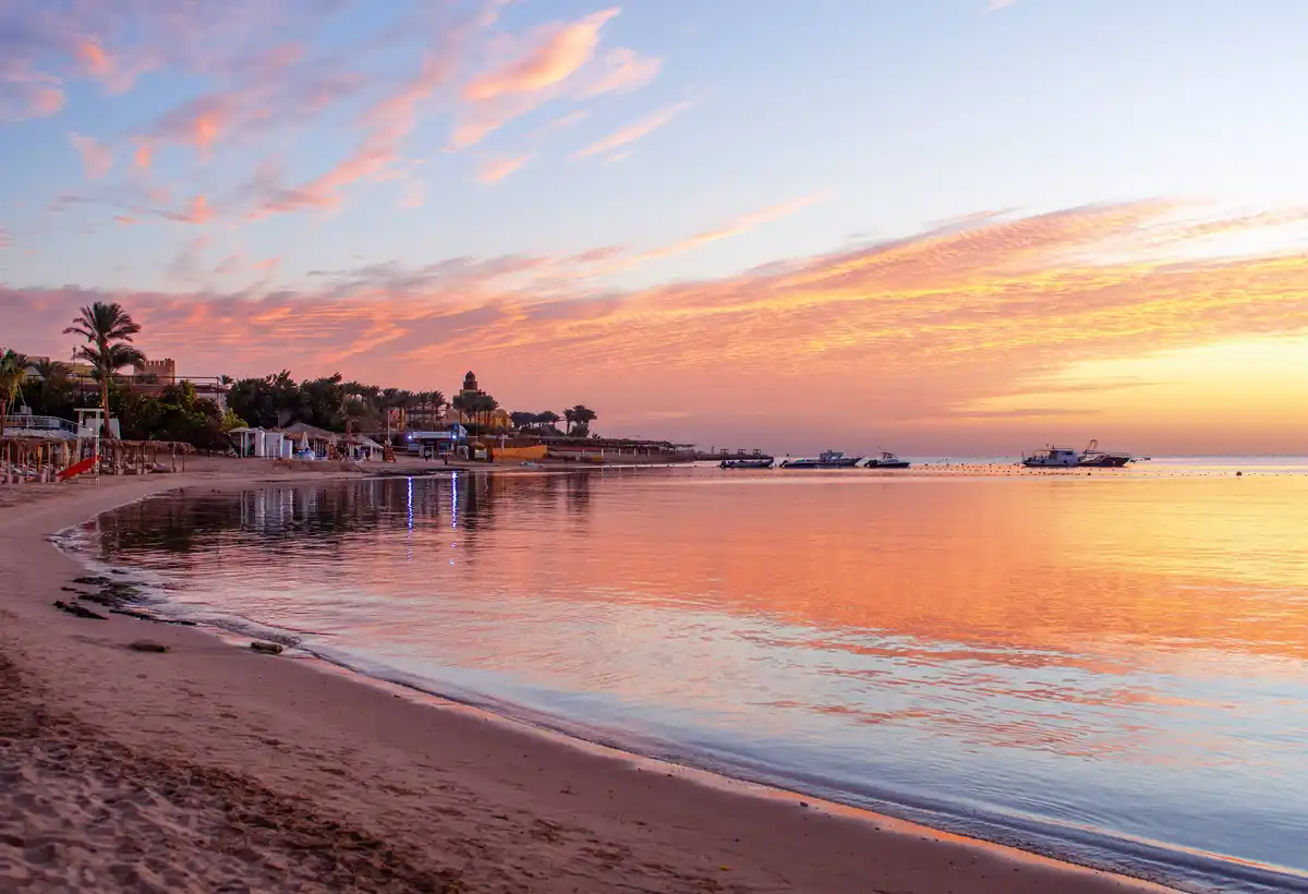 A serene beach at sunset with calm water reflecting orange and pink clouds, palm trees, beach huts, and boats in the…
