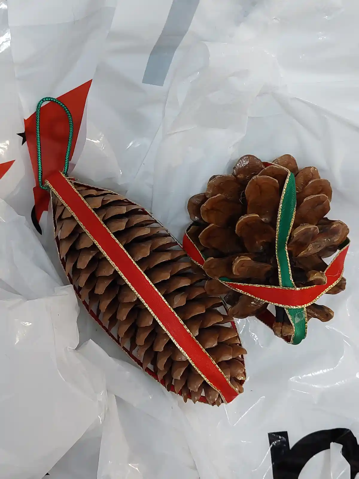 Two pine cone Christmas ornaments, one elongated and one round, are decorated with red and green ribbons with gold trim and…