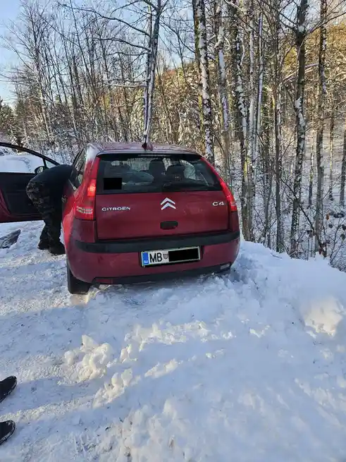 A red Citroën C4 is stuck in deep snow in a forest, with a person in camouflage pants bending over the open driver's side…