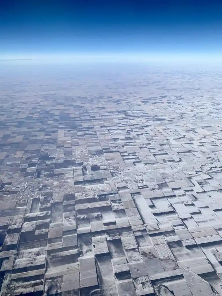 An aerial view from an airplane window shows a vast landscape of patchwork farmland stretching to the horizon under a clear…