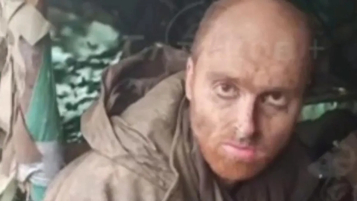A close-up image of a man with a shaved head and dirt on his face, wearing a brown hooded jacket, looking intensely at the…