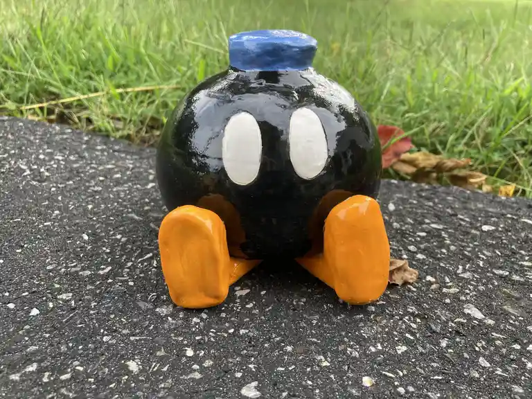 A handmade ceramic figurine resembling a cartoon bomb with a black body, white oval eyes, orange feet, and a blue top sits…