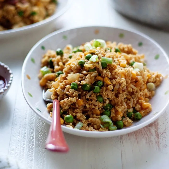 A close-up photo of a bowl of fried rice with peas and green onions, with a pink spoon resting in it. The image has the…