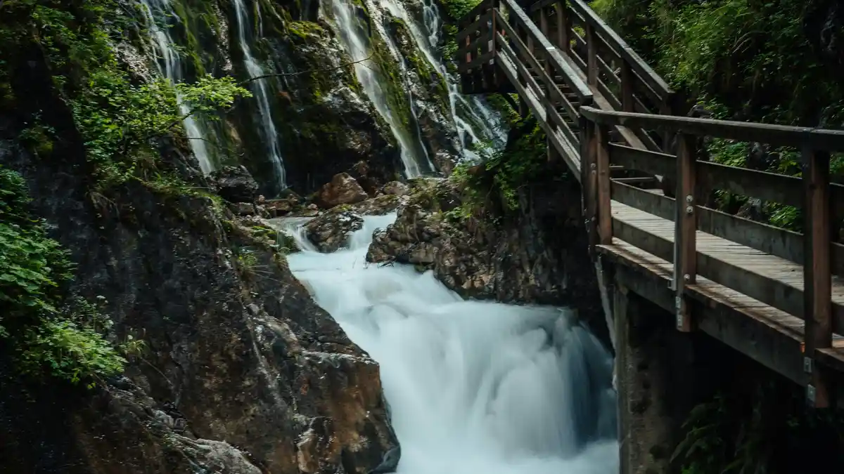 Tumbling waters dance down moss-covered rocks, creating a misty veil that rises to envelop a rustic wooden bridge, its…