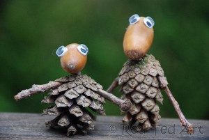 Two handmade pinecone figures with acorn heads and googly eyes stand holding hands on a wooden surface. The pinecone on the…