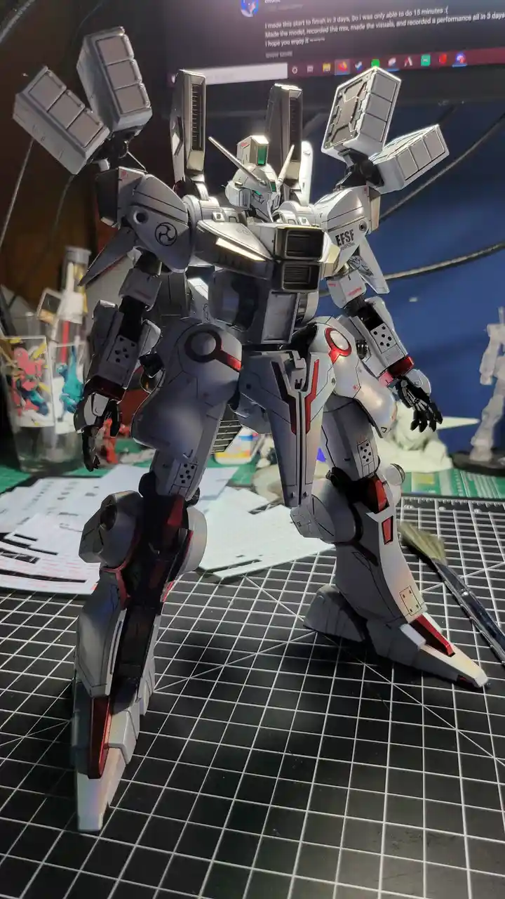 A detailed silver and red Gundam-style robot model stands on a black grid mat, surrounded by hobby supplies like decals and…