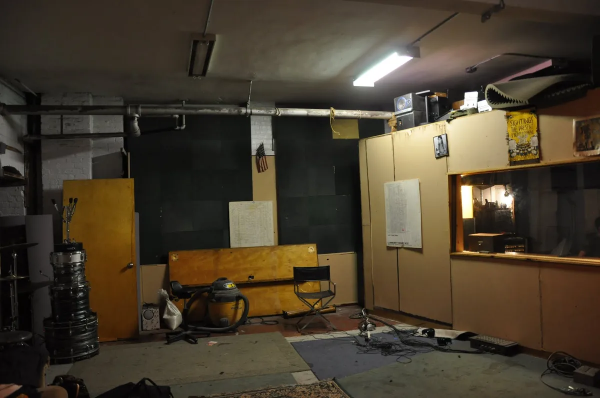 a dimly lit, cluttered basement room with exposed pipes, a stack of drum shells, a vacuum cleaner, a director's chair, and…