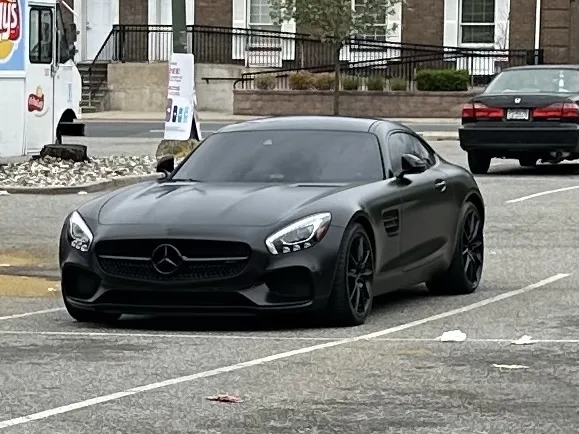 a matte black mercedes-benz gt s sports car is parked in a public asphalt lot, with a white delivery truck and another black…