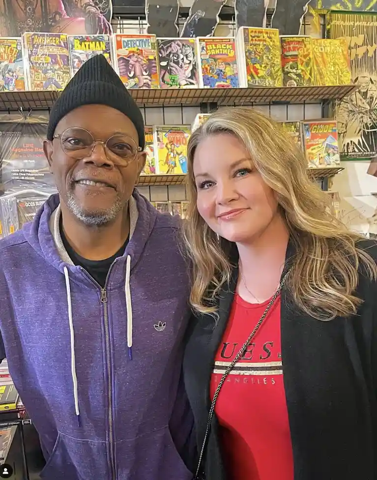 A man and a woman are posing for a photo in a comic book store, with shelves of classic comic books like Avengers, Batman…