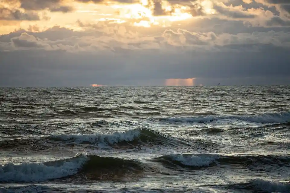 A dramatic seascape at sunset with choppy ocean waves, cloudy sky, and sunbeams breaking through. A lone kitesurfer is…