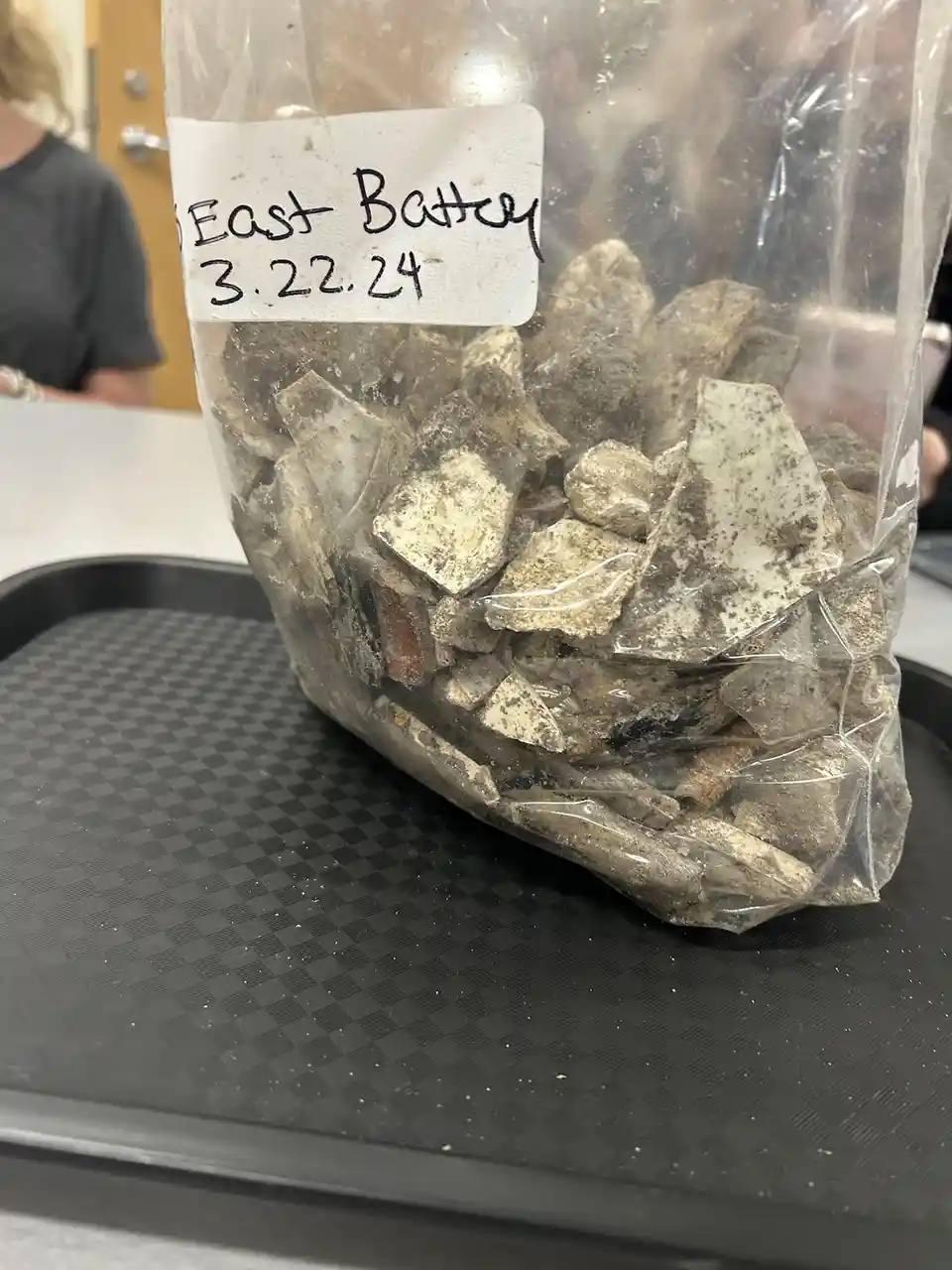 A clear plastic bag filled with dirty, broken ceramic shards sits on a black checkered tray. A handwritten label on the bag…