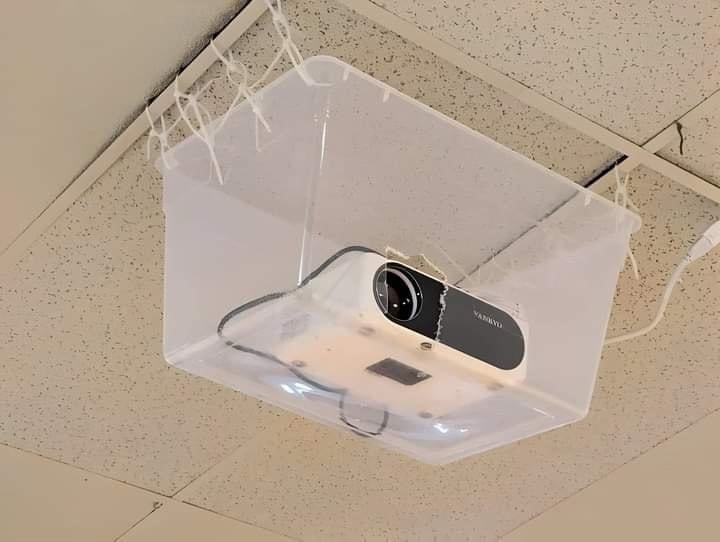 A white and black projector is suspended from a ceiling tile by white zip ties, enclosed within a transparent plastic…