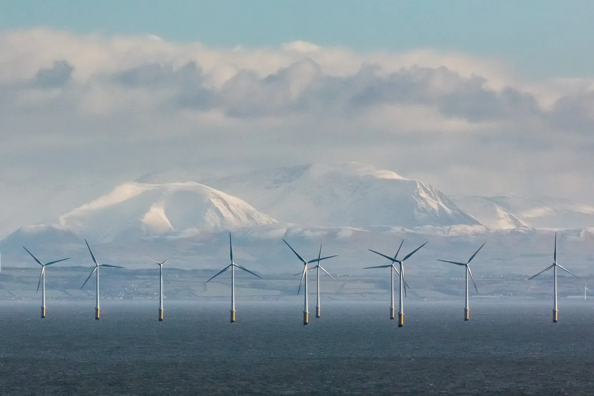A scenic photograph shows multiple offshore wind turbines standing in a body of water, with snow-covered mountains visible…