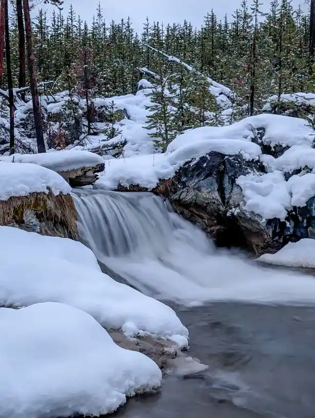 A snowy winter scene shows a small waterfall cascading over rocks covered in thick snow, with evergreen trees in the…