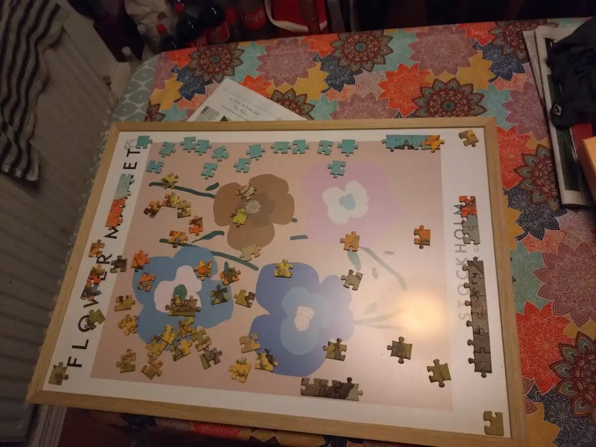 A partially completed jigsaw puzzle of stylized flowers is displayed in a wooden frame on a patterned tablecloth. The…