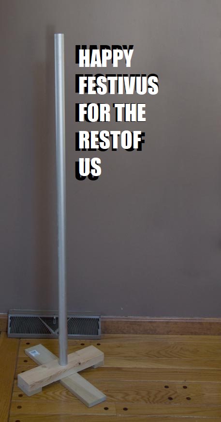 A silver pole stands upright on a wooden floor, mounted on a simple wooden crossbase, against a plain gray wall with a floor…